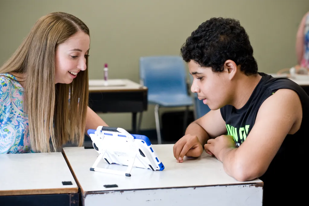 Education student working one-on-one with a middle school student using a tablet on a stand for an instructional activity at a desk.