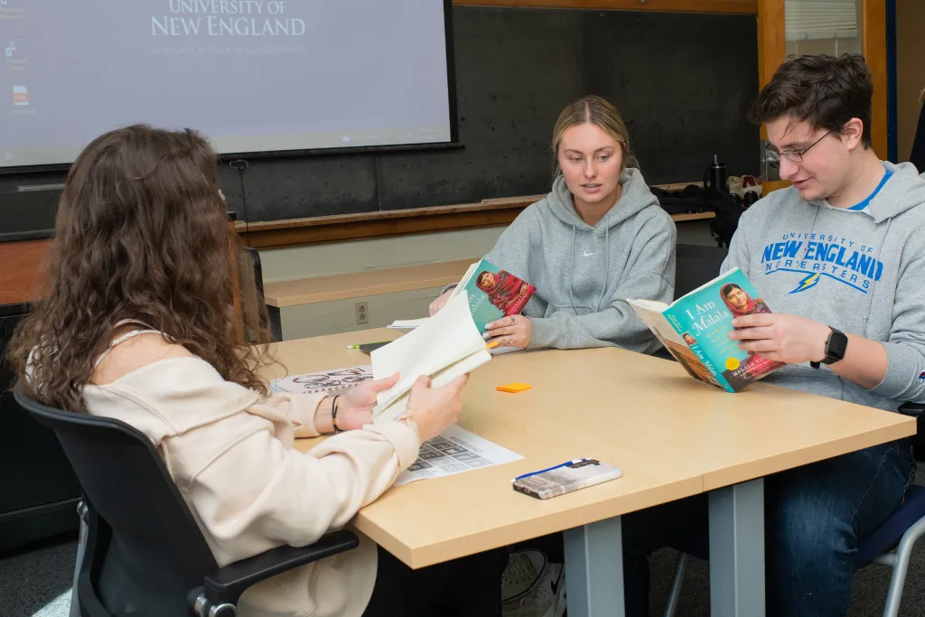 Three students in UNE apparel engaged in a book discussion at a classroom table, with a projection screen displaying the University of New England logo in the background.