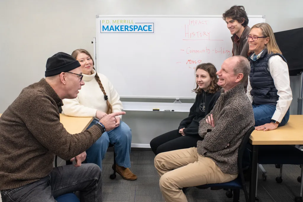 A group of students and U N E staff engage in collaborative discussion in the while seated in a circle around tables with a whiteboard displaying project notes.