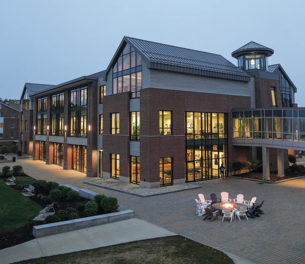 A lit firepit surrounded by Adirondack chairs sits in front of a large brick building with numerous glowing windows at early evening.