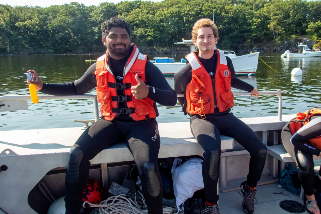 Two marine science students in orange life vests and wetsuits sitting on the side of a research boat, with one giving a thumbs up, during a field study on the water.
