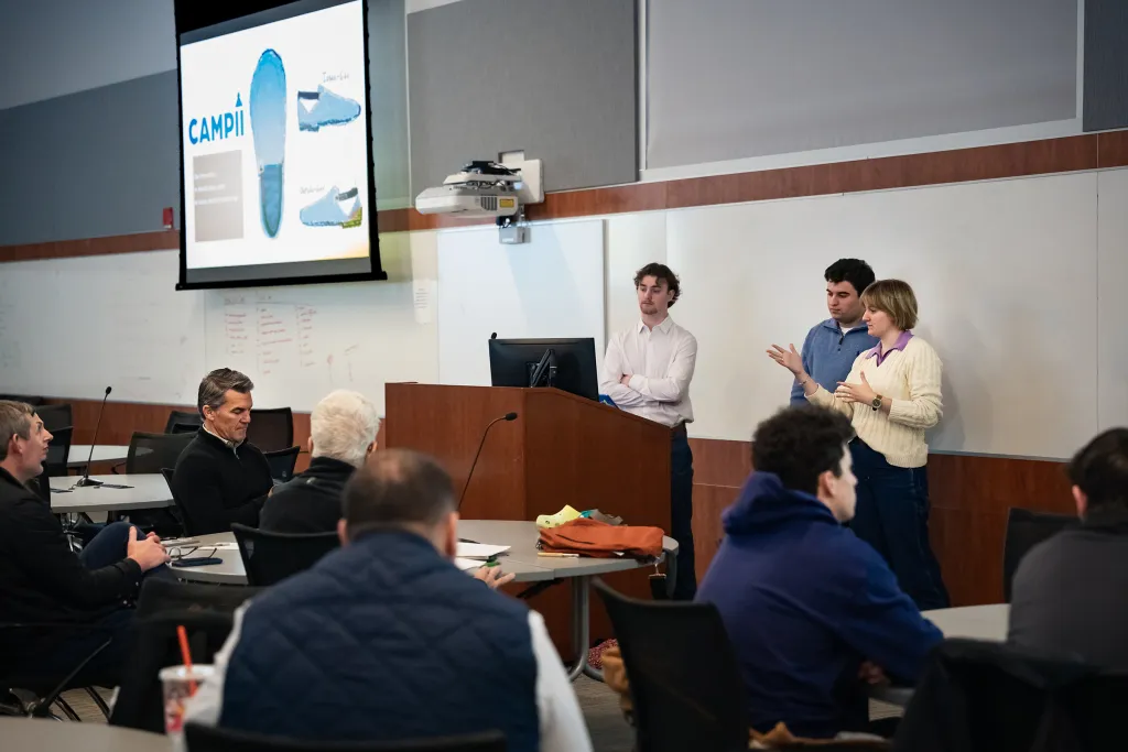 Student team presenting their footwear design project to judges and an audience in a classroom, with a projection screen displaying blue shoe prototypes and product materials on the presentation table.