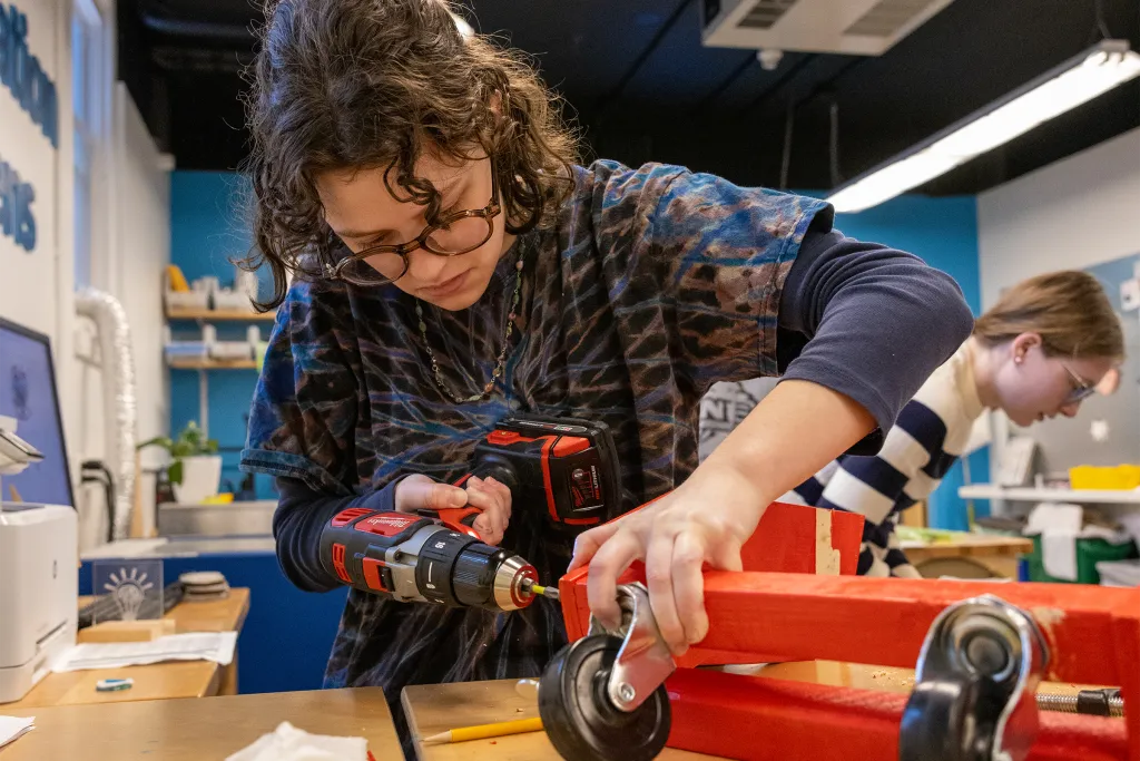 Student in safety glasses using a power drill to assemble a wooden project with wheels in a makerspace workshop.