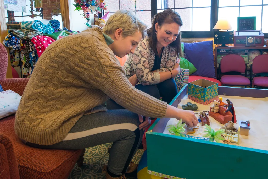 Art therapy session with a U N E social work student and a client working together with miniature figures and sand tray materials in a cozy room with colorful blankets, board games, and large windows.