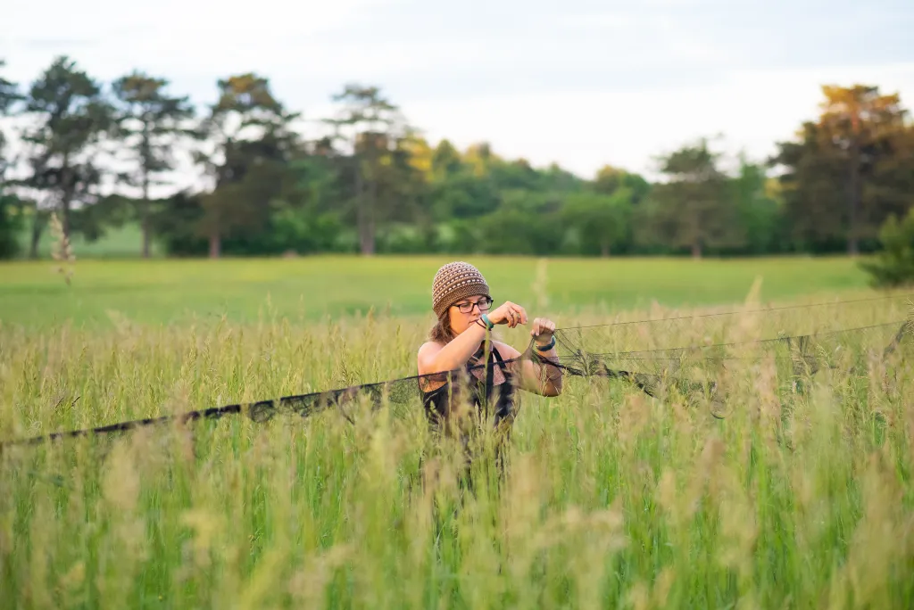 Student conducting field research in tall grass, adjusting equipment on a wire fence with pasture and trees visible in the background.