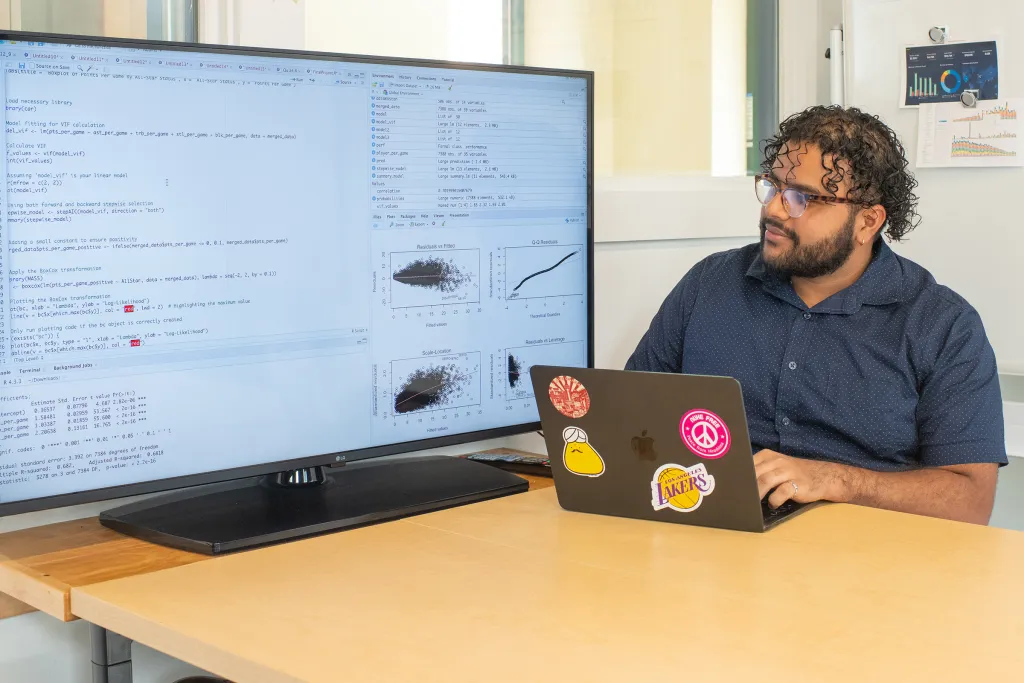 A student analyzes data science code and statistical visualizations on a large monitor while working on a sticker-covered laptop at a desk.