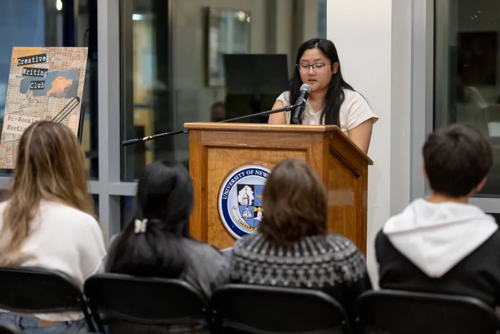 Student presenting at a podium with microphone during a Creative Writing Club event, with audience members seated and a club poster visible in the background.