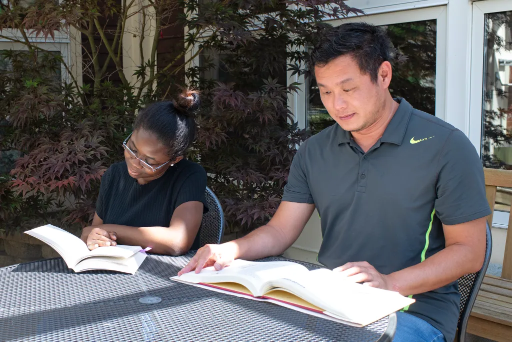 Two students studying together at an outdoor patio table with open textbooks, surrounded by landscaping and building windows in the background.