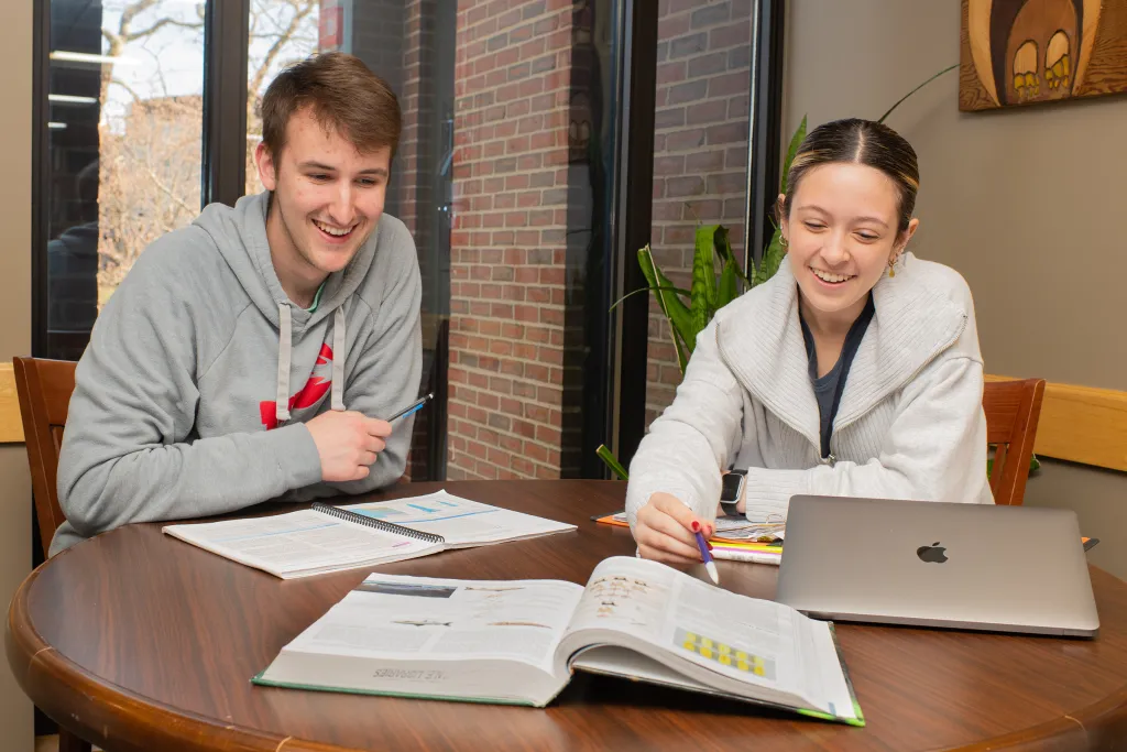 Two students collaborating on coursework at a study table near large windows, with textbooks, notebooks, laptop, and colorful highlighters spread between them.