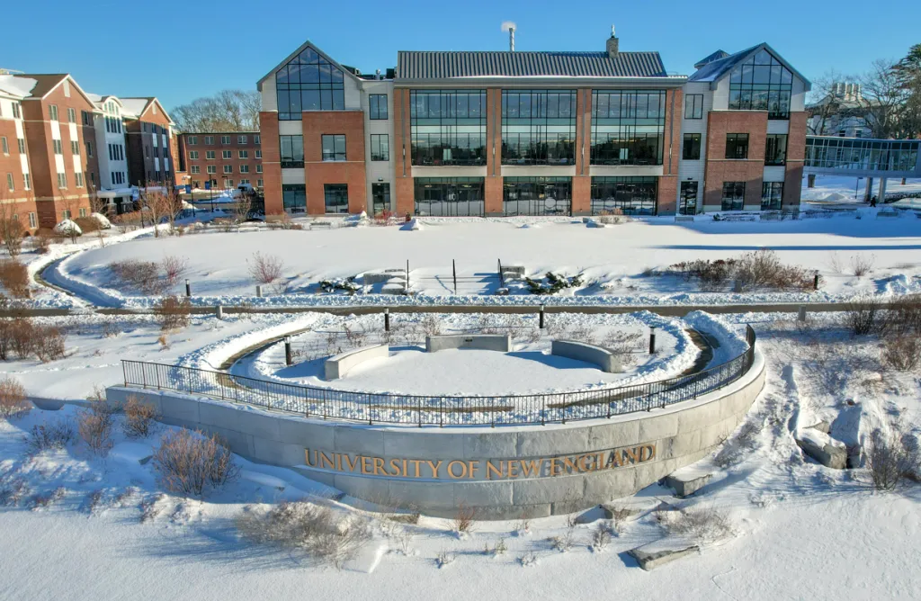 Circular stone amphitheater with 'University of New England' inscription in foreground, modern glass-fronted academic building and residence halls visible across snowy landscape.