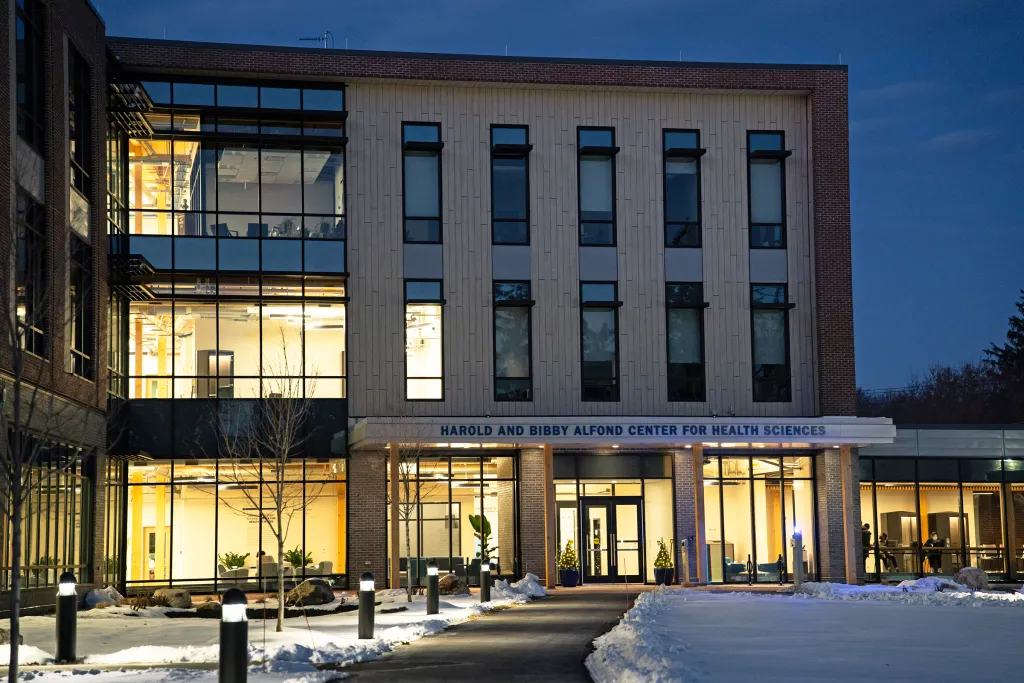 Harold and Bibby Alfond Center for Health Sciences illuminated at dusk in winter, showing modern architecture with large glass windows and snow-covered walkway.