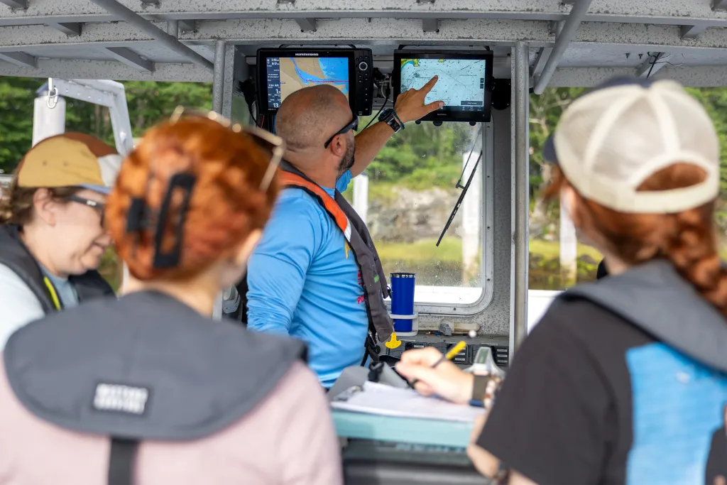 Marine Science instructor points to navigation displays on a boat console while explaining to master's students gathered around during field training.