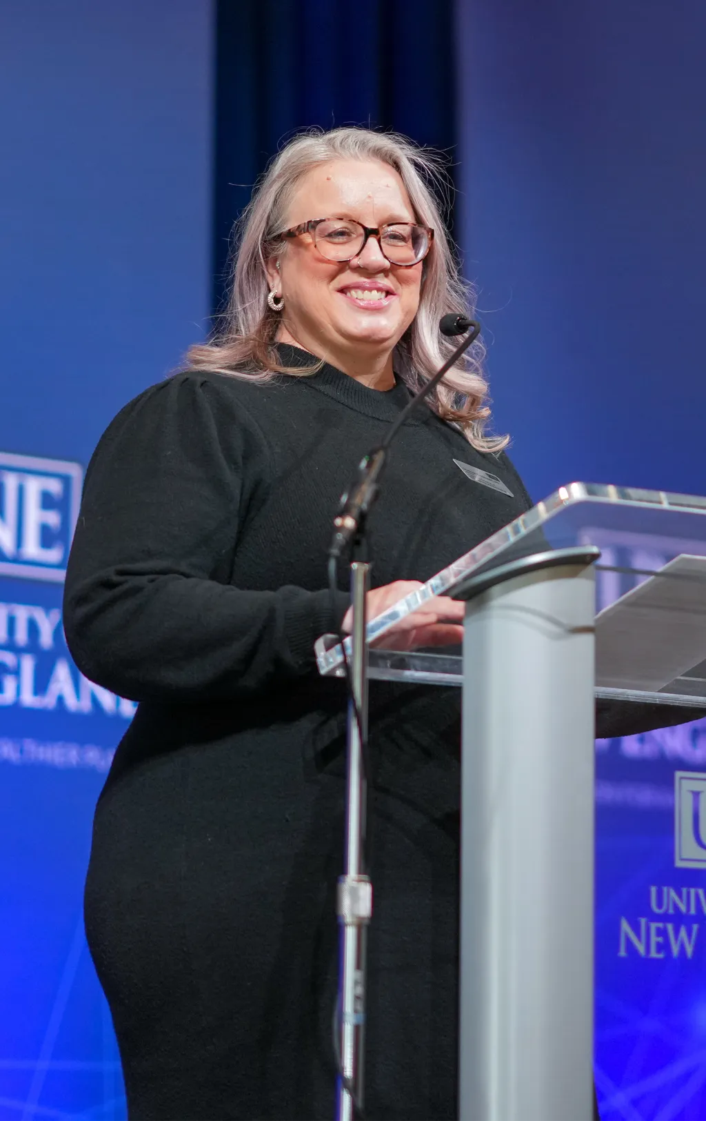 Shannon Zlotkowski stands at podium with microphone delivering presentation at University of New England event with blue branded backdrop.