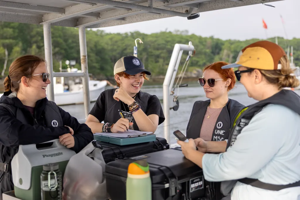 Four graduate marine science students collaborate on a research boat, taking notes and discussing observations during field work.