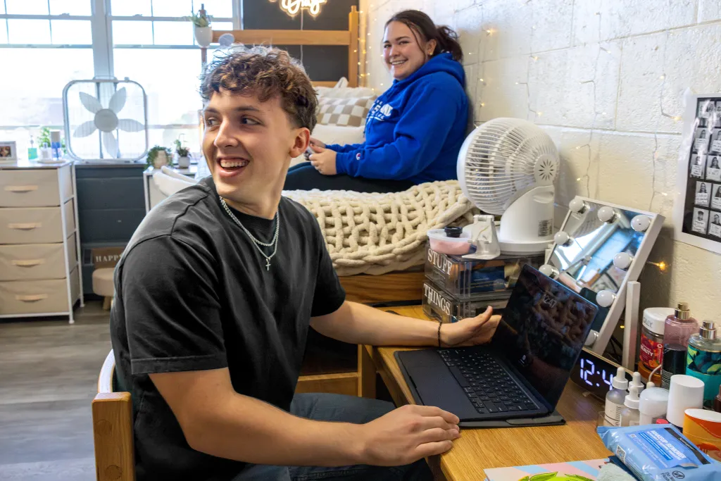 Student works at a laptop at a wooden desk while another student relaxes on a lofted bed behind them in a dorm room with string lights, white brick walls, and personal care items on the desk.