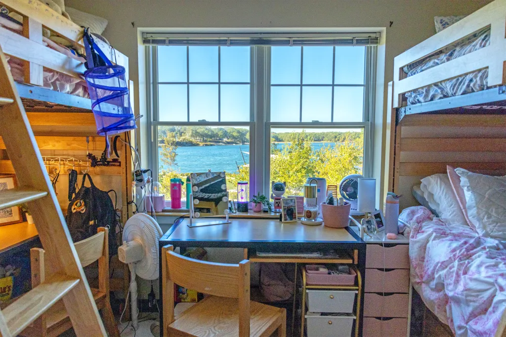 Dorm room with lofted beds on both sides, a central desk facing large windows with a scenic view of a lake and trees, and personal items including water bottles, storage organizers, and a fan.