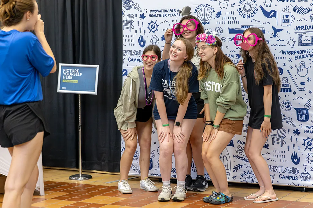 A group of students laugh and pose with fun prop glasses and accessories at a UNE photo booth during an accepted students event, in front of a illustrated backdrop featuring campus landmarks and the phrase "Picture Yourself Here."