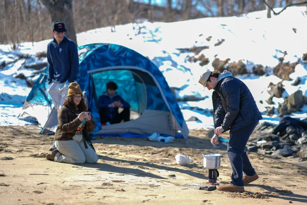 Four students camp on a snowy beach, with one tending a camp stove, another crouching to take a photo, a third looking on, and a fourth sitting inside a blue dome tent.