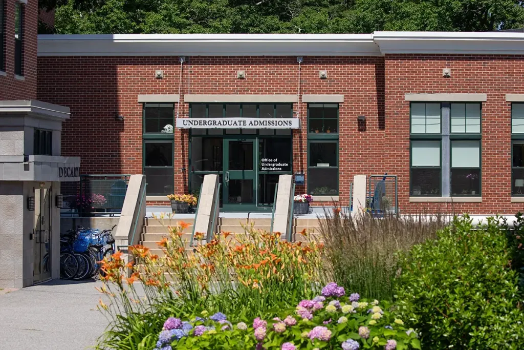 Brick building with "Undergraduate Admissions" sign above glass entrance doors, surrounded by colorful flowering plants and landscaping on campus.