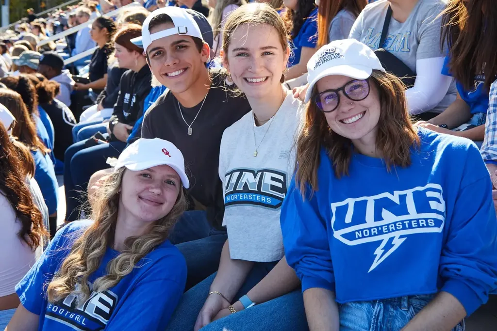 Four smiling students in UNE Nor'easters gear pose together in a packed stadium during a sunny fall football game.
