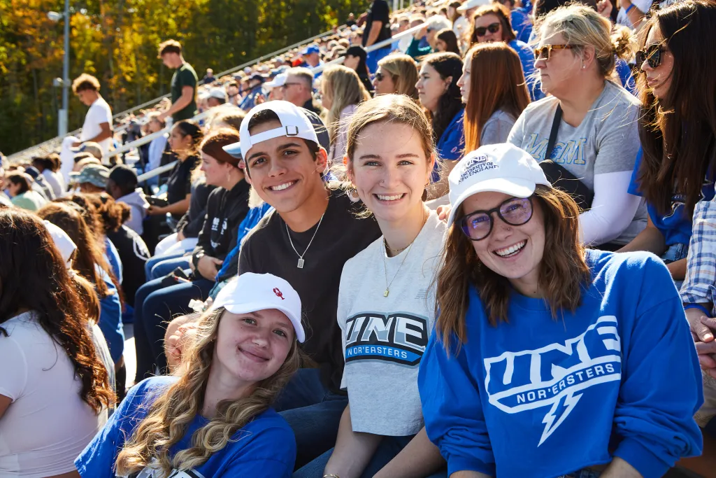 Four smiling students in UNE Nor'easters gear pose together in a packed stadium during a sunny fall football game.