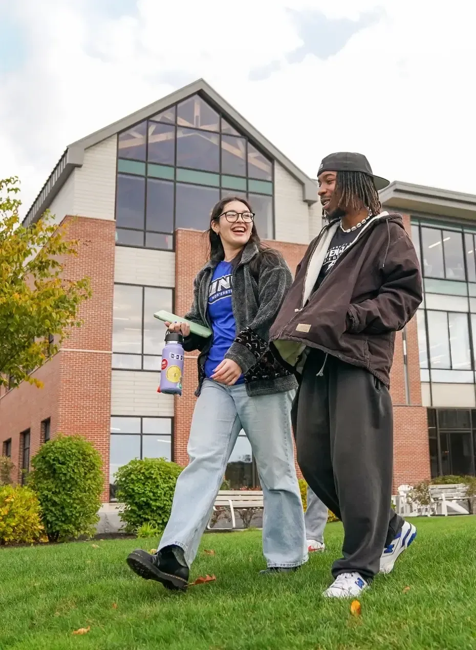 Two students laugh and chat while walking across the grass in front of a brick and glass campus building on a cloudy fall day.