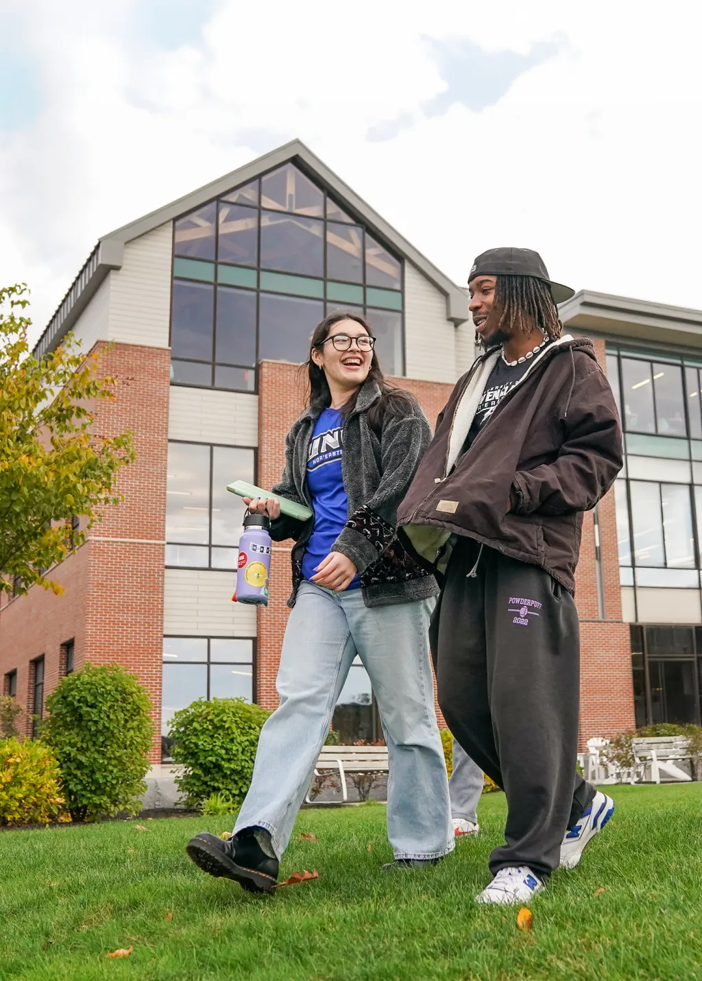 Two students laugh and chat while walking across the grass in front of a brick and glass campus building on a cloudy fall day.