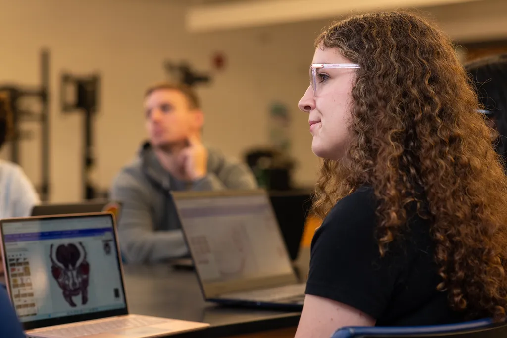 A student with curly hair and glasses attends a clinical anatomy class, with a laptop in the foreground displaying a 3D anatomical model and other students visible in the background.