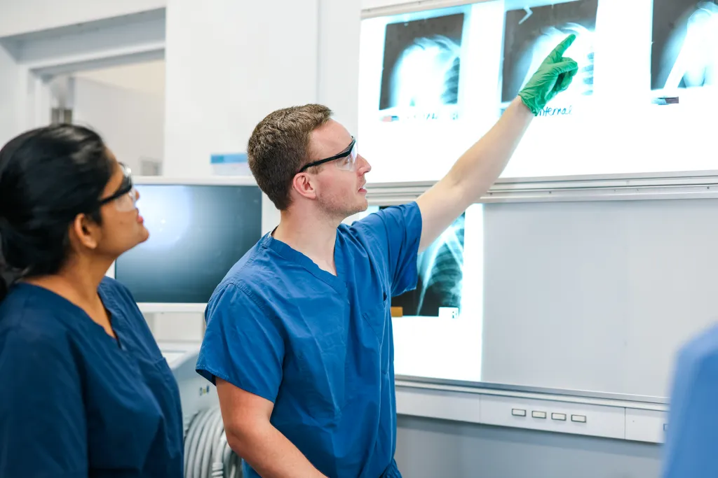 Two students in blue scrubs and safety glasses examine X-ray images on a light board in a clinical anatomy lab, with one student pointing to a chest radiograph.