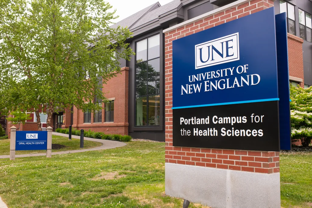 Exterior signage for the UNE Portland Campus for the Health Sciences, with the UNE Oral Health Center sign and a brick building visible in the background surrounded by spring greenery.