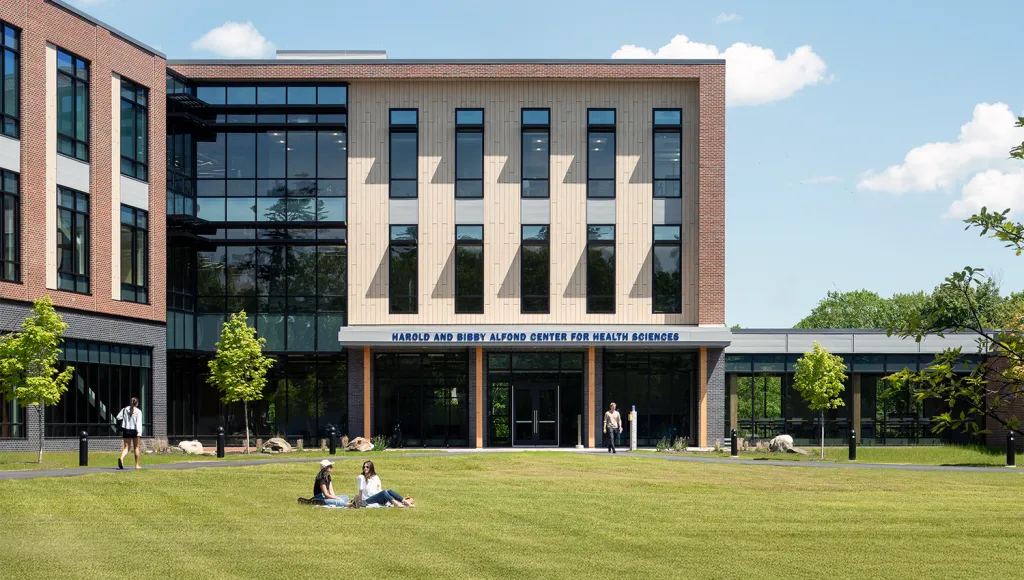 Two students sit on the lawn in front of the Harold and Bibby Alfond Center for Health Sciences on a sunny day, while other students walk along nearby paths.