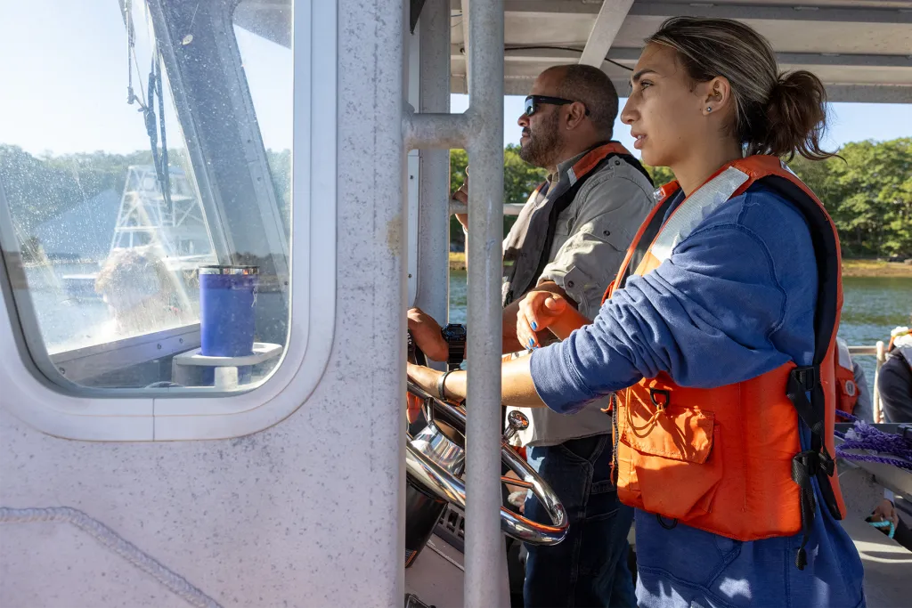 A UNE marine sciences student in an orange life jacket stands at the helm of a research vessel alongside an instructor, both looking forward as the boat moves through the water.