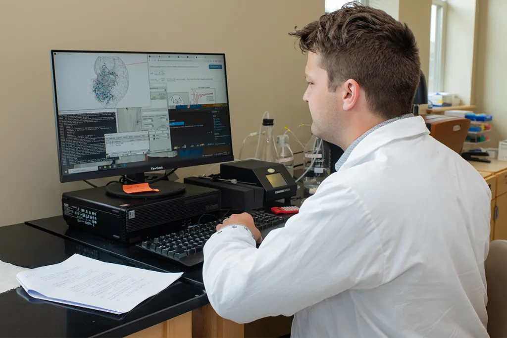 A graduate student in a white lab coat sits at a workstation in a laboratory setting, analyzing complex molecular or network visualization software on a large monitor, with handwritten notes and lab equipment nearby.
