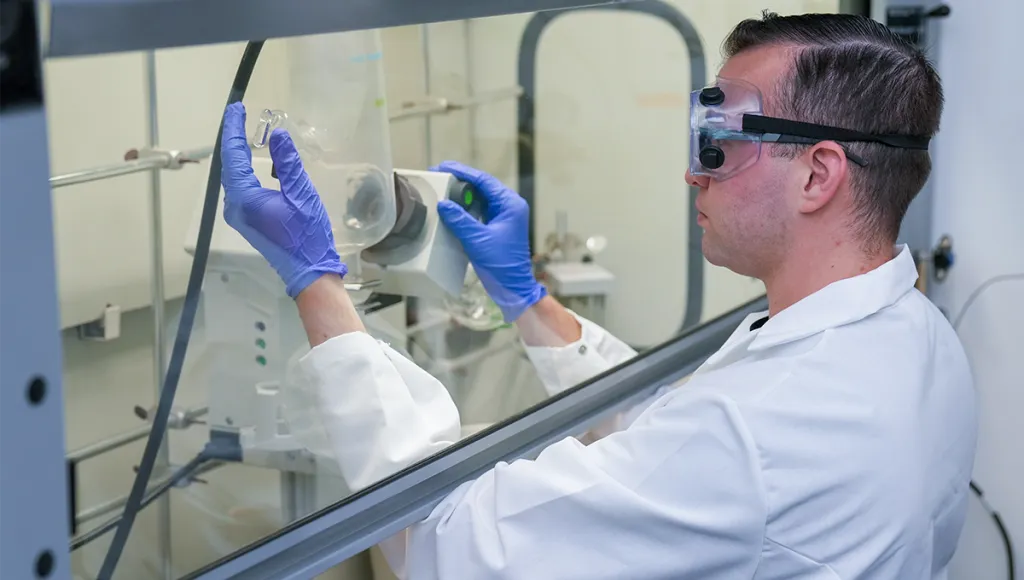 A graduate student in a white lab coat and safety goggles examines a small glass vial while working inside a fume hood, wearing blue nitrile gloves.