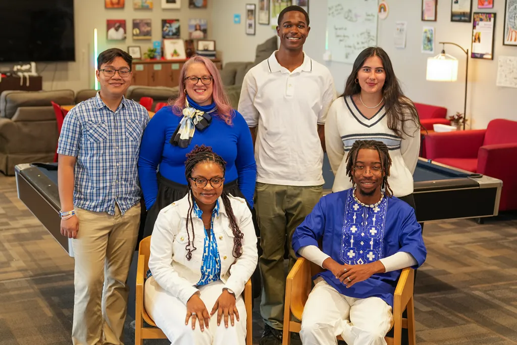A diverse group of six students and one staff member pose together in the UNE Office of Community Belonging, a welcoming lounge space with colorful artwork, a pool table, and comfortable seating visible in the background.