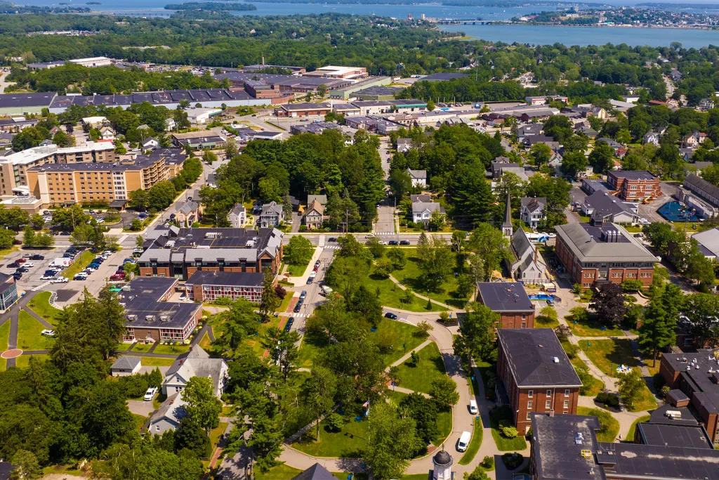 Aerial view of UNE's Portland campus in summer, with tree-lined walkways, historic brick buildings, and Casco Bay visible in the background.
