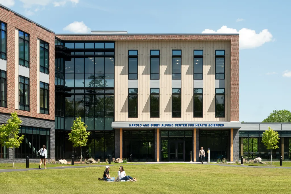 Two students sit on the lawn in front of the Harold and Bibby Alfond Center for Health Sciences on a sunny summer day, with other students walking near the building's entrance.
