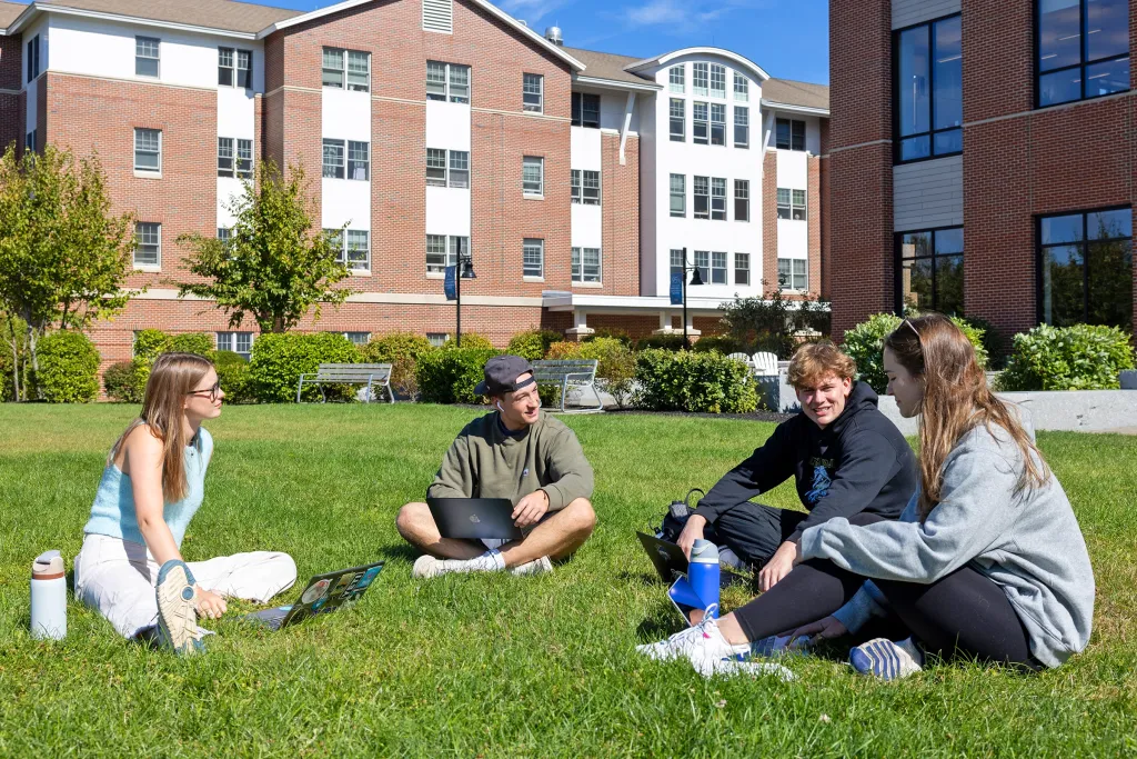 Four students sit together on a sunny campus lawn with laptops, engaged in conversation in front of a brick residence hall.