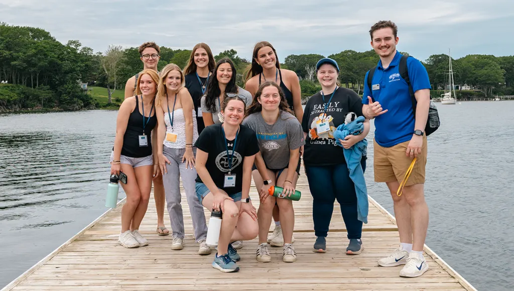 A group of new UNE students and an orientation leader pose together on a waterfront dock during a campus orientation event, with a sailboat and tree-lined cove visible behind them.