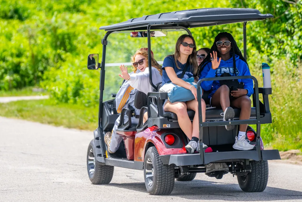 A group of students wave and smile from a golf cart on a tree-lined road on Peaks Island.