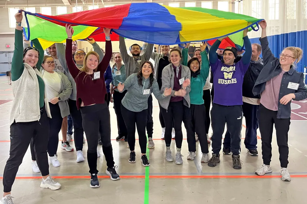 Group of smiling adults wearing name tags raise a colorful parachute overhead together in a gymnasium during a team-building activity.