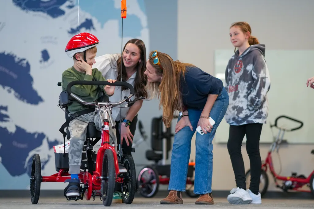 A young boy wearing a red helmet sits on an adaptive tricycle while two smiling women help adjust the fit and a girl looks on nearby.