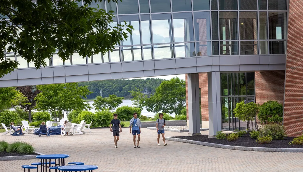 Three students wearing lanyards walk across a sunny brick courtyard beneath a glass pedestrian bridge connecting campus buildings overlooking the water.