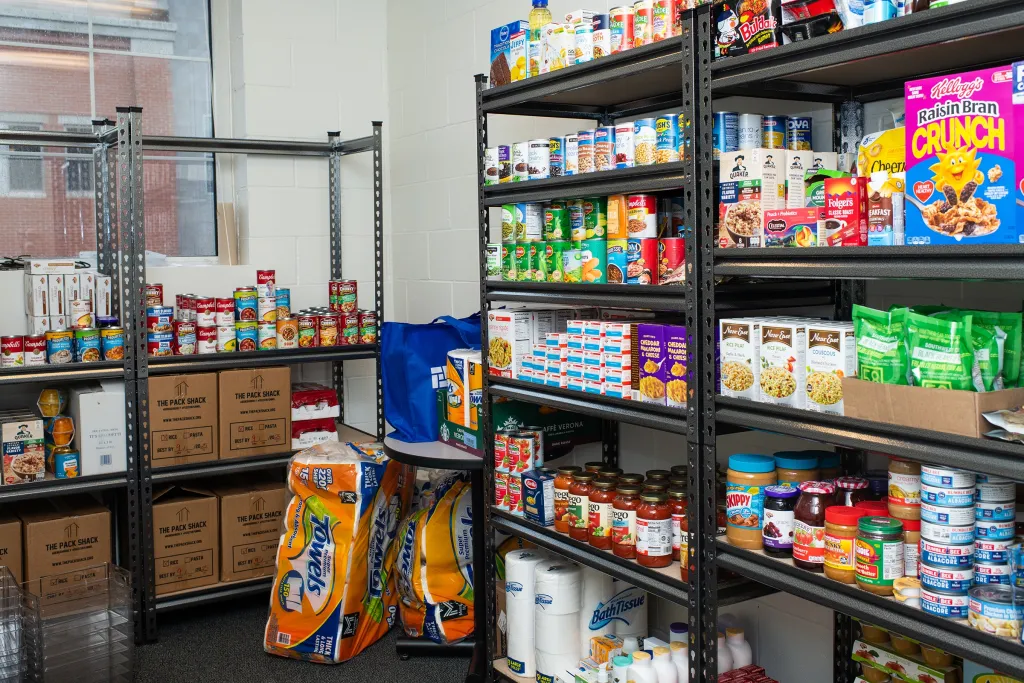 Metal shelves stocked with canned goods, boxed meals, cereals, pasta sauces, peanut butter, and personal care items in a campus food pantry, with bulk paper towels and Pack Shack donation boxes on the floor.