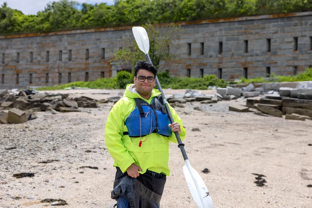 A student in a yellow jacket and blue life jacket holds a kayak paddle on the rocky beach at Fort Gorges, with the fort's granite walls visible behind him.