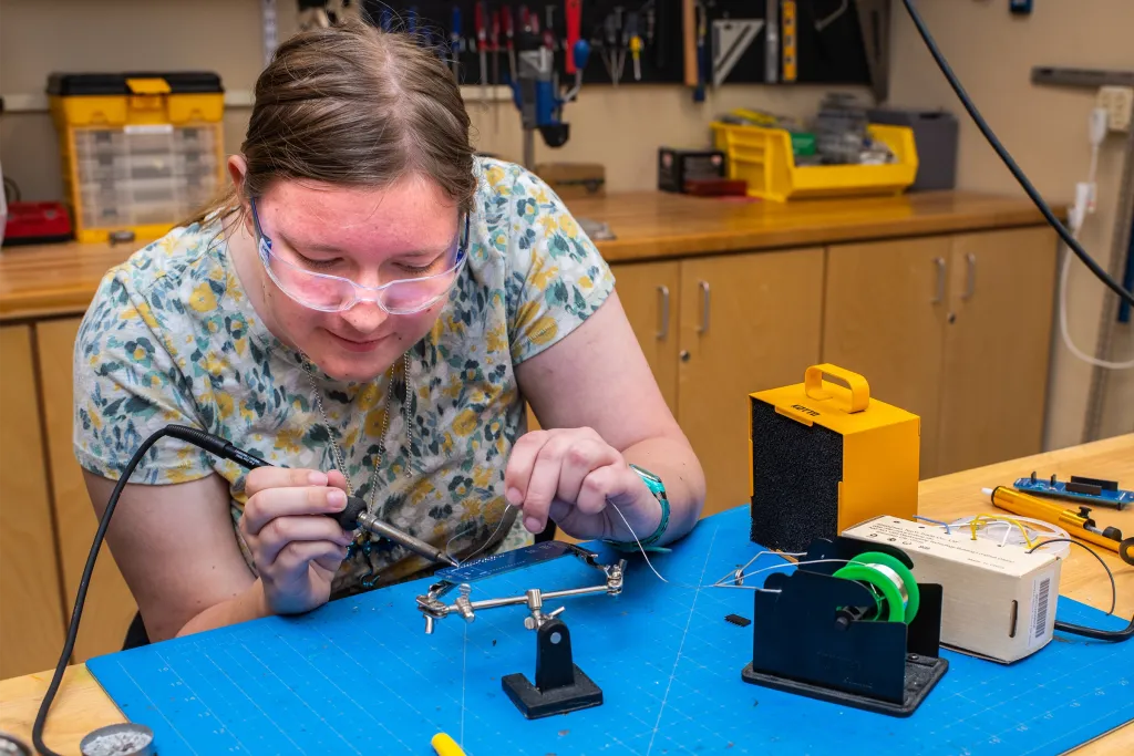 A student wearing safety goggles solders a circuit component on a blue cutting mat in the U N E Makerspace. Tools, spools of solder, and electronic components are arranged on the workbench around her.