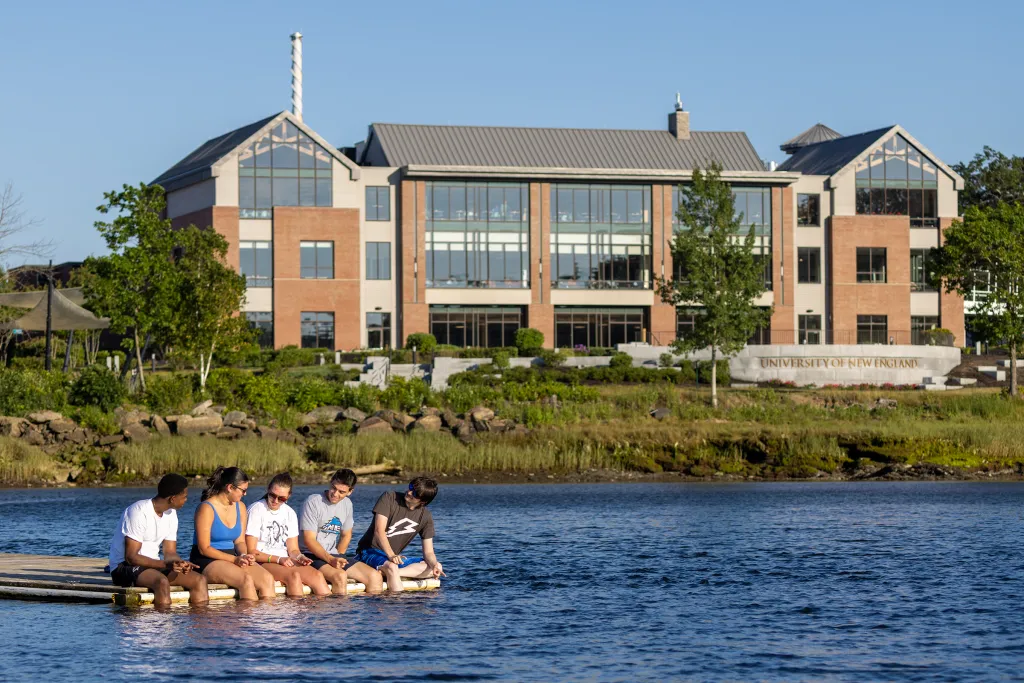 Five students sit on a floating dock with their feet in the water, with a modern three-story brick and glass building with peaked rooflines visible on the shore behind them.