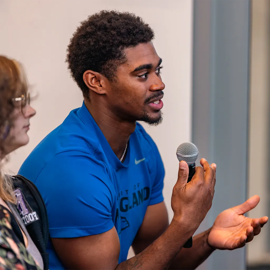 Student in a blue shirt gestures while speaking into a microphone during a classroom discussion.