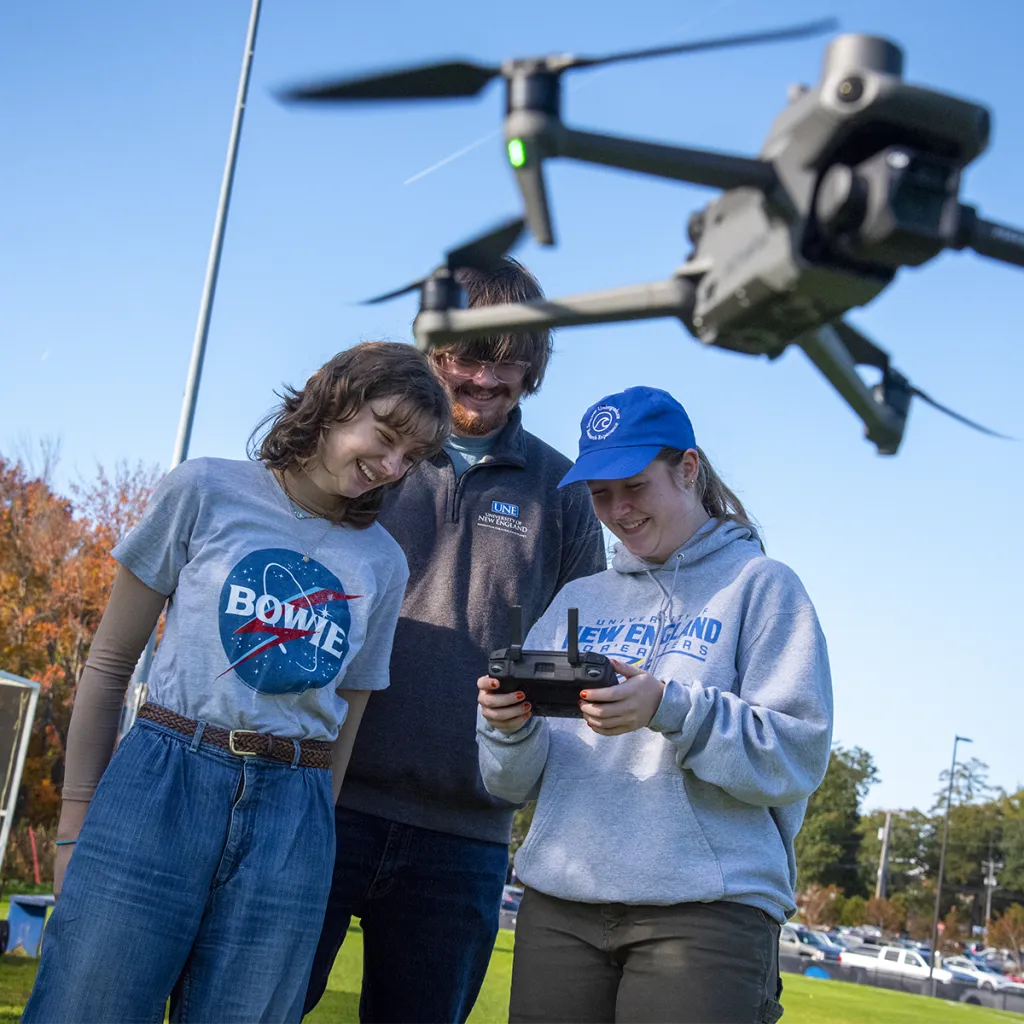 Three students operate a drone outdoors on a sunny autumn day, with one wearing a UNE shirt holding the remote controller while the drone hovers above them.