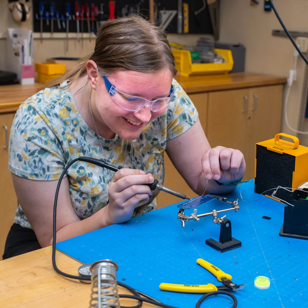 Student wearing safety goggles uses a soldering iron on a small mechanical component at a workbench with blue mat and organized tools.